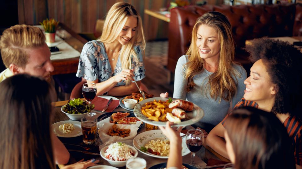 A photograph of some people enjoying some food at a restaurant. They are smiling, holding cutlery and passing plates of food to eachother. 